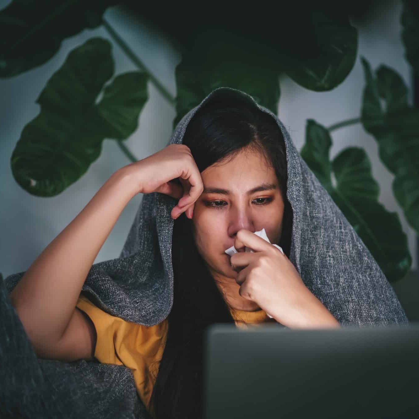 Young asian woman blowing nose and sneeze into tissue paper while working with laptop at night, experiences allergy and flu symptoms, health care concept.